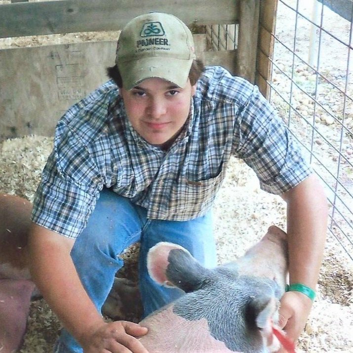 A young farmer in a plaid shirt and cap interacts with a pig in a barn setting, showcasing agricultural practices and animal husbandry. The environment includes wood fencing and straw on the ground, highlighting rural life and farming culture.